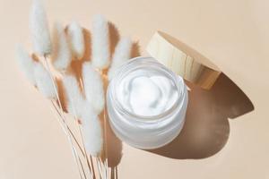 White Moisturizer facial or eye cream in open glass jar and lagurus on beige background. Top view with copy space. photo