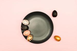 Top view of pink, white and golden decorated eeaster eggs on black plate on pink background. Trendy holiday backdrop. photo