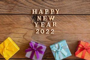 feliz año nuevo 2022. cita hecha de letras de madera y números 2022 sobre fondo de madera con pequeñas cajas de regalo multicolores. concepto creativo para la tarjeta de felicitación de año nuevo foto