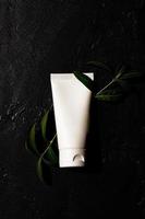 white cosmetic tube with hand cream on a dark background with green leaves. Cosmetics concept with herbal ingredients photo