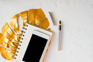 escritorio de mujer. hoja de monstera tropical dorada, bloc de notas en blanco, teléfono inteligente blanco y bolígrafo sobre fondo de hormigón blanco. foto
