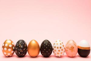 vista lateral de huevos de pascua decorados en rosa, blanco, negro y dorado en una fila sobre fondo rosa. telón de fondo de moda. foto