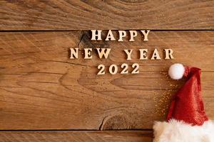 tarjeta de felicitación festiva para el año nuevo 2022 con sombrero rojo de santa y texto de madera en una mesa. espacio de copia para texto foto