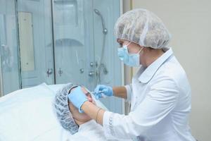 Portrait of  middle aged woman doctor cosmetician in white lab coat, mask and sterile gloves makes injection of Botox into the forehead of a young woman for removing the wrinkles photo