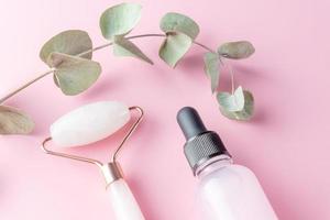 beauty products - facial roller, cosmetic oil and serum on pink background euqalyptus leaves, top view. Natural cosmetics and beauty treatment. photo