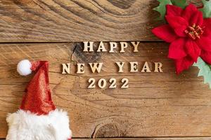 Happy New Year 2022. Quote made from wooden letters and numbers 2022 on wooden background with santa hat and poisentia flower. Creative concept for new year greeting card photo