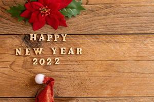 Happy New Year 2022. Quote made from wooden letters and numbers 2022 on wooden background with santa hat and poisentia flower. Creative concept for new year greeting card with copy space photo