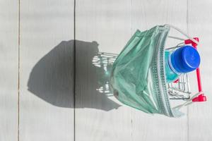 Shopping cart in medical mask with bottle of sanitizer on wgite wooden background. Coronavirus prevention concept. Top view. photo