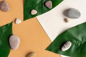 Zen flat lay composition with green leafs and stones. Tropical leaf and sea pebble on beige background. Spa or beauty template with place for text or product . Blank card with tropical leaves. photo