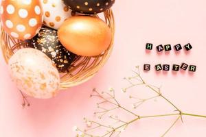 Top view of wooden painted eggs in gold, black and rose colors in wicker basket with branch of gypsophila on pink background. Happy easter text made of black beads photo