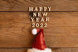 tarjeta de felicitación festiva para el año nuevo 2022 con sombrero rojo de santa y texto de madera en una mesa. copie el espacio para el texto. foto