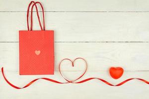 Creative Valentine Day romantic composition with red hearts, satin ribbon and paper bag on wooden background. Mockup with copy space for blogs and social media. photo