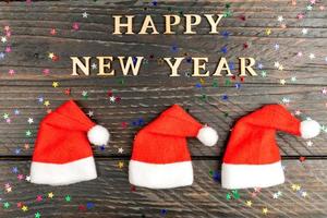 tarjeta de felicitación festiva para feliz año nuevo con texto de madera y hets de santa rojo sobre fondo de madera. foto