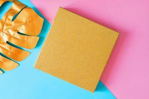 Top view of golden gift box and monstera leaf on double pink and blue background. Minimal flat lay, creative backdrop with copy space photo