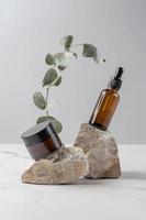 close up of collagen cream in glass jar, serum in amber glass bottle and dry eucalyptus branch on gray background. Set for skin and body care beauty products. photo