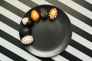 Black and white striped background with white, black and golden decorated eggs on black plate. Trendy easter flat lay. photo
