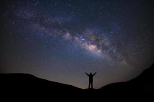 Panorama landscape with milky way, Night sky with stars and silhouette of a standing sporty man with raised up arms on high mountain. photo