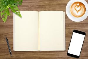 un cuaderno en blanco abierto, un teléfono inteligente con bolígrafo y una taza de café con leche en una mesa de madera. foto