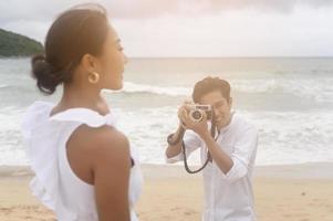 Happy young couple wearing white dress enjoy taking photo on the beach on holidays, travel, romantic, wedding concept