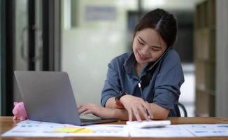 Portrait of Asian young female talking on the phone with a business customer while using a calculator recording data on her work computer. photo