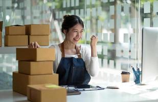 Startup SME small business entrepreneur of freelance Asian woman using a laptop with box Cheerful success Asian woman her hand lifts up online marketing packaging box and delivery SME idea concept photo