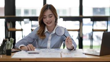 joven empresaria asiática que trabaja en una computadora portátil con una calculadora y examina un contrato meticulosamente con una lupa en la oficina foto