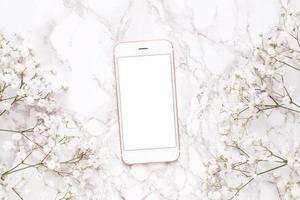 Mobile phone mock up and white flowers on marble table top view in flat lay style. Woman working desk . photo