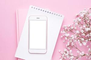 Mobile phone mock up and white flowers on pink pastel table top view in flat lay style. Woman working desk. photo