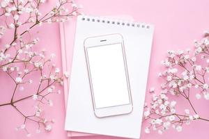 Mobile phone mock up and white flowers on pink pastel table top view in flat lay style. Woman working desk. photo