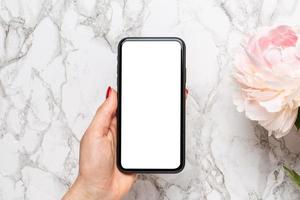 Mobile phone in in female hand with piony flowers on a marble background, using technology concept for the holidays - Valentine's Day, Women's and Mother's Day. Floristics and decoration photo