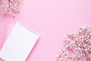 Empty notebook and white flowers on pink pastel table top view in flat lay style. Woman working desk. photo