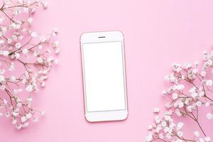 Mobile phone mock up and white flowers on pink pastel table top view in flat lay style. Woman working desk. photo