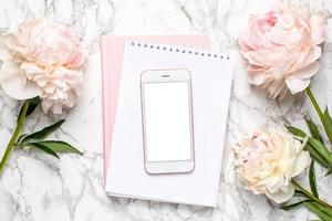 Mobile phone with a white and pink notebook and piony flowers on a marble background photo