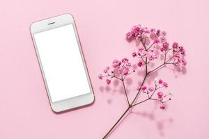 Mobile phone mock up and pink flowers on pink pastel table top view in flat lay style. Woman working desk.Spring colour photo