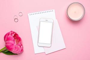 Mobile phone mock up, notebook and peony flower on pink pastel table in flat lay style. Woman working desk.Summer colour photo