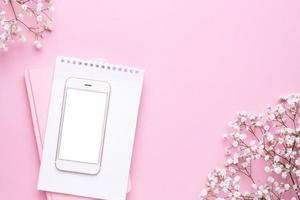 Mobile phone mock up and white flowers on pink pastel table top view in flat lay style. Woman working desk. photo