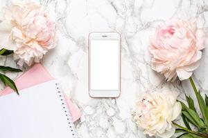 Mobile phone with a white and pink notebook and piony flowers on a marble background photo