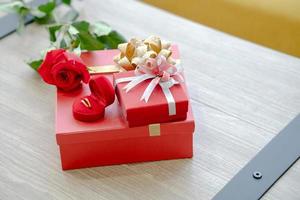 caja de regalo con cinta roja, anillo de oro, flor rosa en la mesa de madera foto