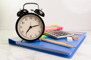 black clock with 7 am 12 minutes with blue folder and writing equipment with Calculator white table at the office photo