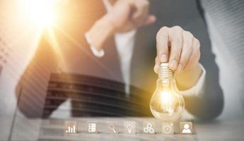 Businessman hand holding light bulb with icon on wood cube and   working on desk, Creativity and innovation are keys to success.Concept of new idea and innovation with Brain and light bulbs, photo