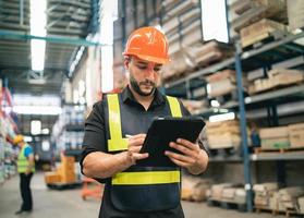 Professional manager man employee using tablet check stock working at warehouse. Worker wearing high visibility clothing and a hard hat, helmet and checking and count up goods or boxes for delivery. photo