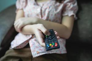 cierre el control remoto de la televisión en las manos de una joven camisa rosa apuntando al televisor y encendiéndolo o apagándolo. seleccione el canal viendo la televisión en el sofá de su casa en la sala de estar, relájese. foto