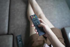 cierre el control remoto de la televisión en las manos de una joven camisa rosa apuntando al televisor y encendiéndolo o apagándolo. seleccione el canal viendo la televisión en el sofá de su casa en la sala de estar, relájese. foto