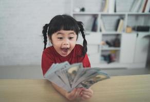 Asian baby girl wearing a red t-shirt holding dollar bill on wood table desk in livingroom at home. Saving investment wealth concept. photo