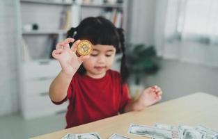 Asian baby girl wearing a red t-shirt holding gold bitcoin crypto currency blockchain on wood table desk in livingroom at home. Crypto currency blockchain Saving investment wealth concept. photo