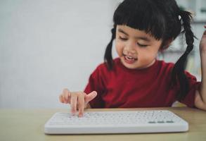 una niña asiática que usa una camiseta roja usa un teclado inalámbrico y estudia en línea en un escritorio de madera en la sala de estar en casa. educación aprendiendo en línea desde el concepto de hogar. foto