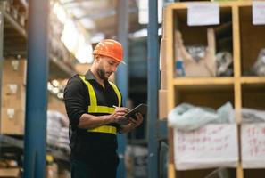 Professional manager man employee using tablet check stock working at warehouse. Worker wearing high visibility clothing and a hard hat, helmet and checking and count up goods or boxes for delivery. photo