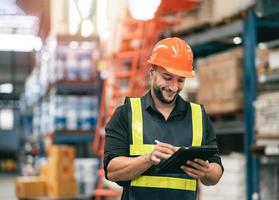 Professional manager man employee using tablet check stock working at warehouse. Worker wearing high visibility clothing and a hard hat, helmet and checking and count up goods or boxes for delivery. photo