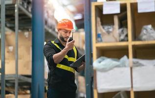 Manager or engineer man employee use walkie-talkie to give an order to co-worker and checking stock at warehouse. Worker wearing high visibility clothing, helmet count up goods or boxes for delivery. photo