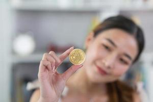 Asian woman girl smiling holding gold bitcoin crypto currency blockchain on wood table desk in living room at home. Crypto currency blockchain Saving investment wealth concept. photo